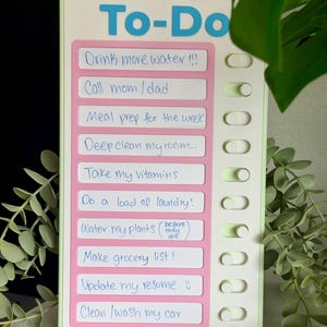May include: A rectangular white and light green plastic to-do list board with a pink center panel. It features the blue text To-Do at the top and ten white horizontal slots with handwritten tasks and corresponding sliding toggles on the right side.