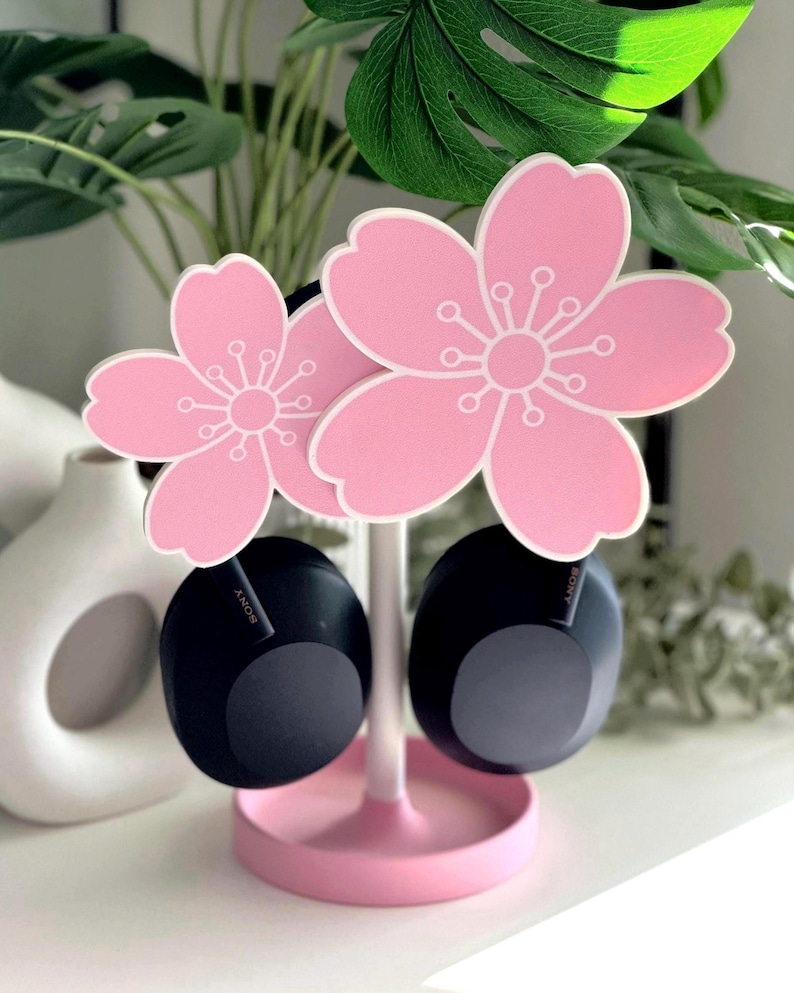 Op de afbeelding: Een roze hoofdtelefoonstandaard in de vorm van kersenbloesems. De standaard houdt twee zwarte hoofdtelefoons vast. De standaard heeft een ronde basis en een witte steel. De kersenbloesems zijn roze met witte details.