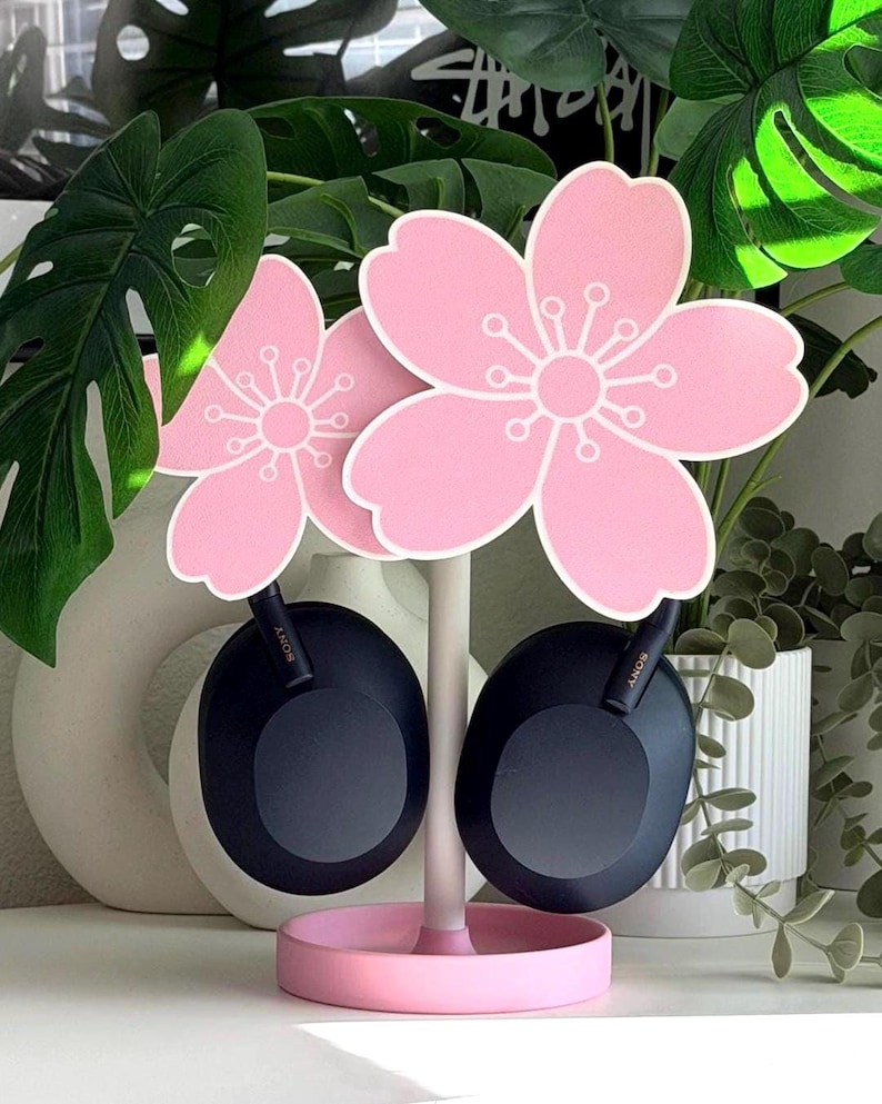 Op de afbeelding: Een roze hoofdtelefoonstandaard in de vorm van kersenbloesems, met zwarte hoofdtelefoons. De standaard heeft een ronde basis en een witte steel. Het kersenbloesem-ontwerp is roze met witte details. De hoofdtelefoons zijn zwart met het merk "SONY".