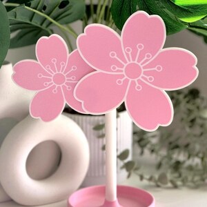 Op de afbeelding: Twee roze kersenbloesemdecoraties met witte accenten. De bloemen zijn gemonteerd op een witte steel en geplaatst in een roze cirkelvormige basis. De bloesems hebben een gedetailleerd wit centrum en bloemblaadjesontwerp. Decoratieve bloemaccenten.