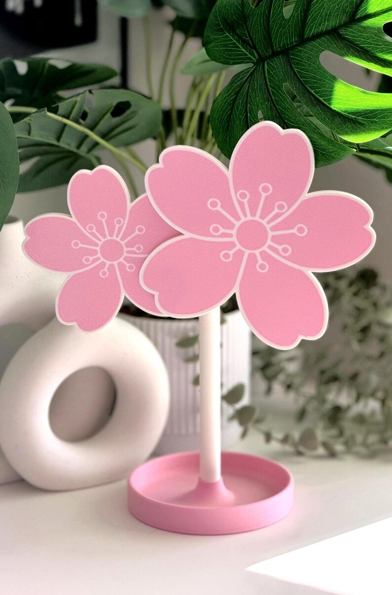 Op de afbeelding: Twee roze kersenbloesemdecoraties met witte accenten. De bloemen zijn gemonteerd op een witte steel en geplaatst in een roze cirkelvormige basis. De bloesems hebben een gedetailleerd wit centrum en bloemblaadjesontwerp. Decoratieve bloemaccenten.