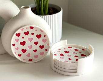 May include: White ceramic coasters with a red and pink heart pattern. The coasters are displayed in a white holder, with a matching coaster resting against a white vase. A decorative plant in a white pot is in the background.