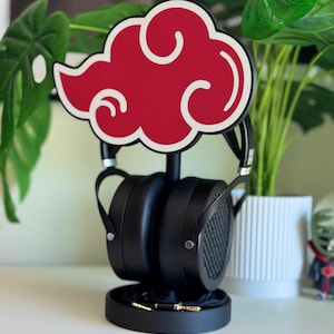May include: Black headphones resting on a black stand, topped with a red cloud-shaped accent with white trim. The stand has a circular base and holds the headphones upright. A cable with gold-colored connectors is visible.