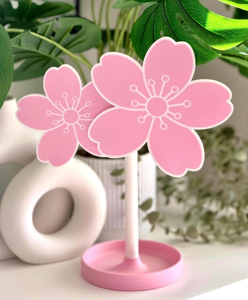 Op de afbeelding: Een roze kersenbloesemdecoratie met witte accenten. De bloem is gemonteerd op een witte steel en staat in een roze cirkelvormige basis. De achtergrond bevat groen en een witte keramische vaas.