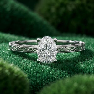 May include: A silver engagement ring with an oval-cut diamond. The band features an intricate, engraved design. The ring is set against a green background, highlighting the sparkle of the diamond.
