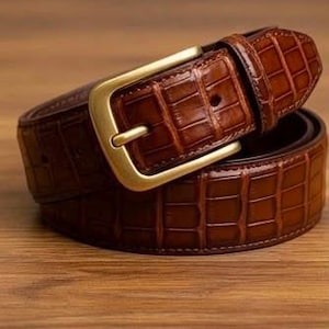 Puede incluir: Un cinturón marrón con una textura brillante en relieve de cocodrilo y una hebilla rectangular de metal en tono dorado cepillado. El cinturón cuenta con una trabilla a juego y una serie de agujeros perforados para su ajuste.