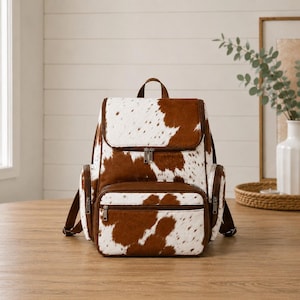 Puede incluir: Una mochila con estampado de piel de vaca en marrón y blanco, que cuenta con una solapa superior, un bolsillo frontal con cremallera y dos bolsillos laterales. Incluye ribetes de piel sintética marrón, cremalleras plateadas y correas ajustables.