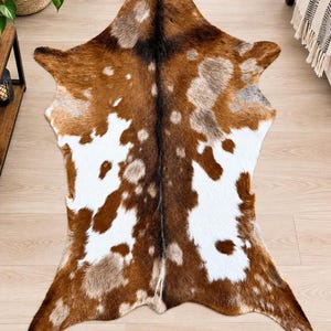 Op de afbeelding: Een koeienhuid vloerkleed met een natuurlijk patroon van bruine, witte en grijze vlekken. De huid heeft een opvallende donkerbruine centrale streep die verticaal door het midden loopt, met onregelmatige witte en lichtbruine vlekken verspreid.