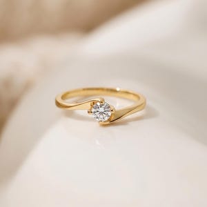 Puede incluir: Un anillo de oro con una sola piedra preciosa transparente de talla brillante redonda engastada en una banda de estilo bypass. La banda de metal tiene un acabado liso y pulido que se curva alrededor de la piedra central.