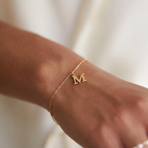 Puede incluir: Una pulsera dorada con una cadena delicada y un colgante con la letra "M". La pulsera se lleva en la muñeca, y la letra "M" destaca en el diseño.
