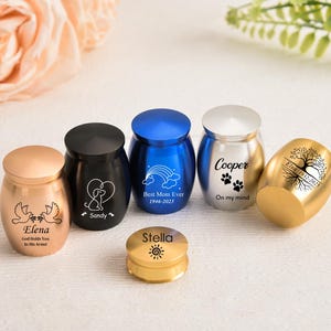 Puede incluir: Una colección de pequeñas urnas coloridas con tapas. Las urnas son de varios colores, incluyendo dorado, negro, azul y plateado. Algunas urnas tienen inscripciones, como "Elena", "Sandy" y "Cooper". Otras tienen elementos decorativos.