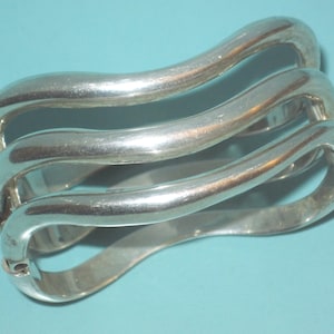 May include: A set of three silver-toned, curved bangles. Each bangle has a smooth, polished surface and a unique, flowing shape. The bangles are connected and appear to be made of metal. The background is a solid light blue.