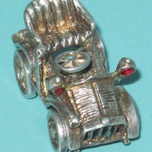 May include: A detailed silver-toned miniature car with a vintage design. The car features a detailed interior, a steering wheel, and red accents on the headlights. The wheels are visible, and the overall design suggests a classic automobile.