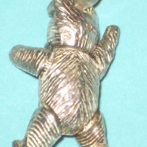 K&ouml;nnte beinhalten: Ein silberfarbener Teddyb&auml;r-Anh&auml;nger mit einer Schlaufe f&uuml;r eine Halskette oder ein Armband. Der B&auml;r wird in einer Laufpose mit strukturierten Details an K&ouml;rper, Armen und Beinen dargestellt. Der Anh&auml;nger ist aus gl&auml;nzendem Metall gefertigt.