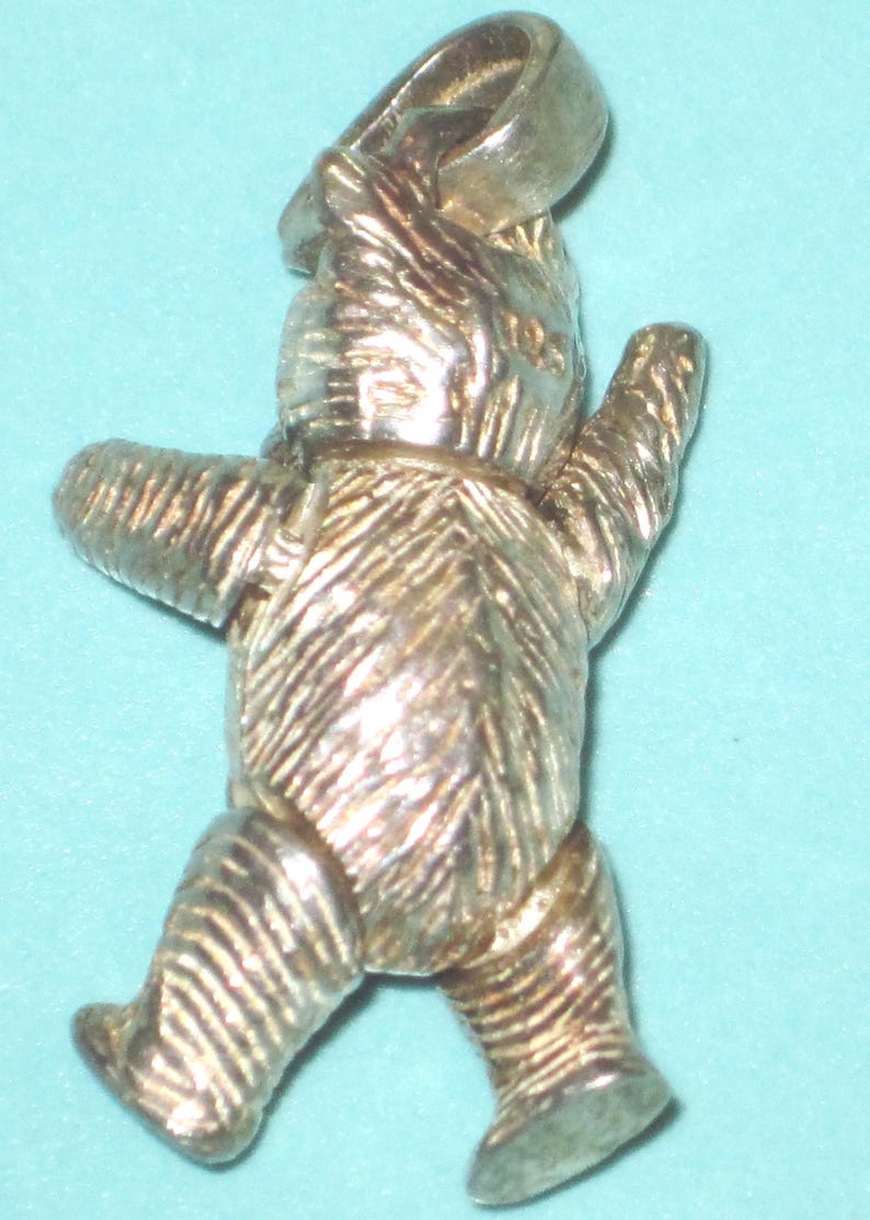 K&ouml;nnte beinhalten: Ein silberfarbener Teddyb&auml;r-Anh&auml;nger mit einer Schlaufe f&uuml;r eine Halskette oder ein Armband. Der B&auml;r wird in einer Laufpose mit strukturierten Details an K&ouml;rper, Armen und Beinen dargestellt. Der Anh&auml;nger ist aus gl&auml;nzendem Metall gefertigt.
