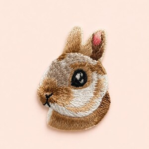 May include: An embroidered patch featuring the head of a brown and white rabbit. The rabbit has a dark eye, a small black nose, and one ear with a pink inner detail. The embroidery uses shades of tan, brown, white, and black thread.