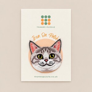 May include: An embroidered iron-on patch featuring a grey and white tabby cat face with yellow eyes, pink ears, and a pink nose. The patch is mounted on a white display card with the text Thames Puzzle and Iron On Patch printed on it.