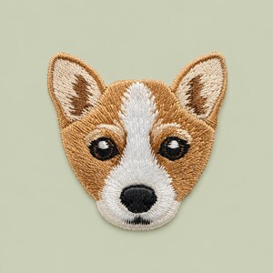 May include: An embroidered patch featuring the head of a Corgi dog. The design displays tan and white fur patterns, large pointed ears with brown accents, dark eyes, and a black nose, all rendered with detailed stitching on a fabric base.