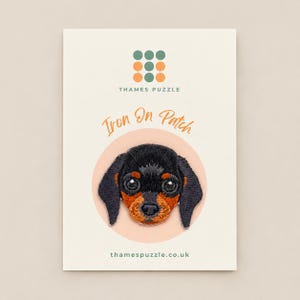 May include: An embroidered iron-on patch featuring a black and tan dachshund puppy face. The patch is displayed on a cream-coloured card with the text Thames Puzzle, Iron On Patch, and thamespuzzle.co.uk printed in green and orange.