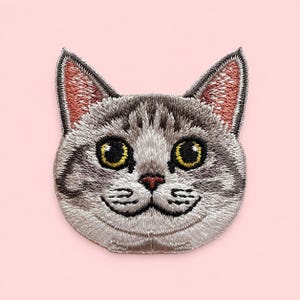 May include: An embroidered iron-on patch featuring the head of a grey tabby cat with dark grey stripes, large yellow eyes, a small pink nose, and pink-toned inner ears. The cat face is detailed with white highlights and black stitching.