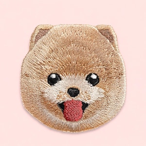 May include: A circular embroidered patch featuring the face of a tan Pomeranian dog. The design includes detailed tan fur stitching, two black eyes with white highlights, a small black nose, and a bright red tongue sticking out of its mouth.