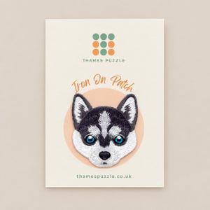 May include: An embroidered iron-on patch featuring a black and white husky puppy face with bright blue eyes. The patch is displayed on a cream-coloured card with the text Thames Puzzle, Iron On Patch, and thamespuzzle.co.uk printed on it.