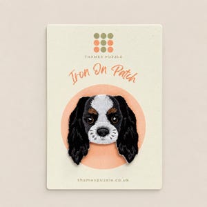 May include: An embroidered iron-on patch featuring a Cavalier King Charles Spaniel dog face with black, white, and tan fur. The patch is mounted on a cream-coloured card labelled with the text Thames Puzzle and Iron On Patch.