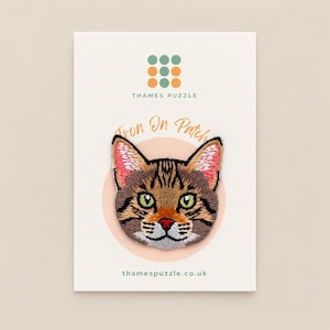 May include: An embroidered iron-on patch featuring a brown tabby cat face with green eyes, pink inner ears, and a red nose. The patch is displayed on a white backing card with the text Thames Puzzle, Iron On Patch, and thamespuzzle.co.uk printed on it.