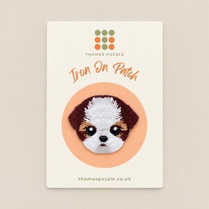 May include: An embroidered iron-on patch featuring a stylised puppy face with white, brown and tan fur, large black eyes and a small black nose. The patch is mounted on a cream-coloured card labelled Thames Puzzle with the text Iron On Patch.