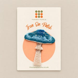 May include: An embroidered iron-on patch featuring a blue mushroom with a white stem, displayed on a rectangular card with the text Thames Puzzle, Iron On Patch, and thamespuzzle.co.uk. The patch has a textured blue cap and a light beige stem.