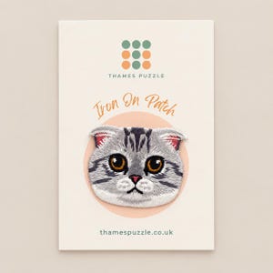 May include: An embroidered iron-on patch featuring a grey and white tabby cat face with large amber eyes and folded ears. The patch is mounted on a cream-coloured card labelled with the text Thames Puzzle, Iron On Patch, and thamespuzzle.co.uk.