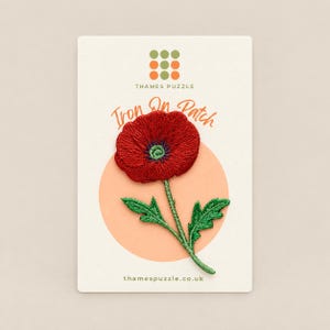May include: An embroidered iron-on patch featuring a vibrant red poppy flower with a green centre and stem with two leaves. The patch is mounted on a rectangular card labelled Thames Puzzle with the text Iron On Patch and thamespuzzle.co.uk.