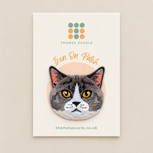 May include: An embroidered iron-on patch featuring a grey and white cat face with bright orange eyes and pink inner ears. The patch is mounted on a cream-coloured card labelled Thames Puzzle Iron On Patch with a grid of green and orange dots.