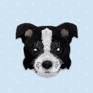 Op de afbeelding: Een geborduurde patch met de kop van een hond met zwarte vacht op de oren en de zijkanten van het gezicht, en een witte streep die van het voorhoofd naar de snuit loopt. De hond heeft donkere ogen en een zwarte neus, uitgevoerd in gedetailleerd stikwerk.