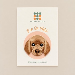 May include: An embroidered iron-on patch featuring a brown puppy face with dark eyes and a black nose. The patch is displayed on a white card with the text Thames Puzzle, Iron On Patch, and thamespuzzle.co.uk printed in black and orange.