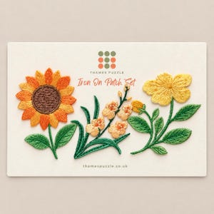 May include: An iron-on patch set featuring three embroidered floral designs on a white card. The patches include a large orange and yellow sunflower, a cluster of small peach-coloured blossoms with green stems, and a bright yellow flower with green leaves.