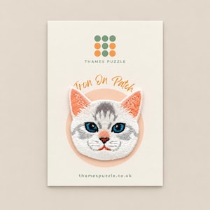 May include: An embroidered iron-on patch featuring a white cat face with grey tabby markings, bright blue eyes, and coral-coloured inner ears. The patch is mounted on a cream-coloured display card with the text Thames Puzzle and thamespuzzle.co.uk.