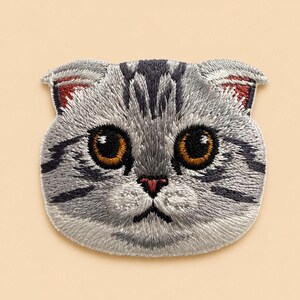 以下が含まれることがあります： 耳が折れた猫の頭の形をした刺繍ワッペン。グレーと白の毛並みにダークグレーの縞模様があり、大きな琥珀色の目、小さなピンクの鼻、耳の内側の赤茶色のディテールが、細かな糸で表現されています。
