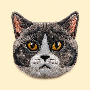 May include: An embroidered patch featuring the head of a cat with grey and white fur, bright amber eyes, a black nose, and pinkish inner ears. The design captures the feline's facial features with detailed stitching in shades of grey, white, and gold.