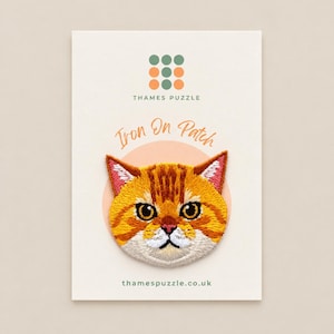 May include: An embroidered iron-on patch featuring a ginger tabby cat face with orange, yellow, and white fur, large dark eyes, and pink inner ears. The patch is mounted on a white display card with the text Thames Puzzle and thamespuzzle.co.uk.