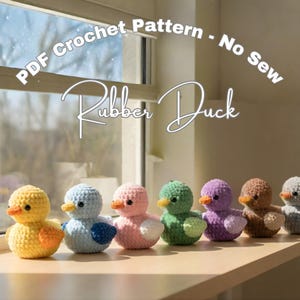 May include: A row of seven colorful crocheted rubber ducks. Each duck has a different color, including yellow, blue, pink, green, purple, brown, and gray. The ducks are displayed on a wooden surface near a window. The text "PDF Crochet Pattern - No Sew" and "Rubber Duck" is visible.