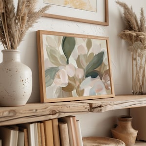 May include: A framed abstract floral painting in a wooden frame. The artwork features soft, muted colours including shades of green, pink, and beige. The painting is displayed on a rustic wooden shelf, alongside a vase with dried plants and a ceramic vase.