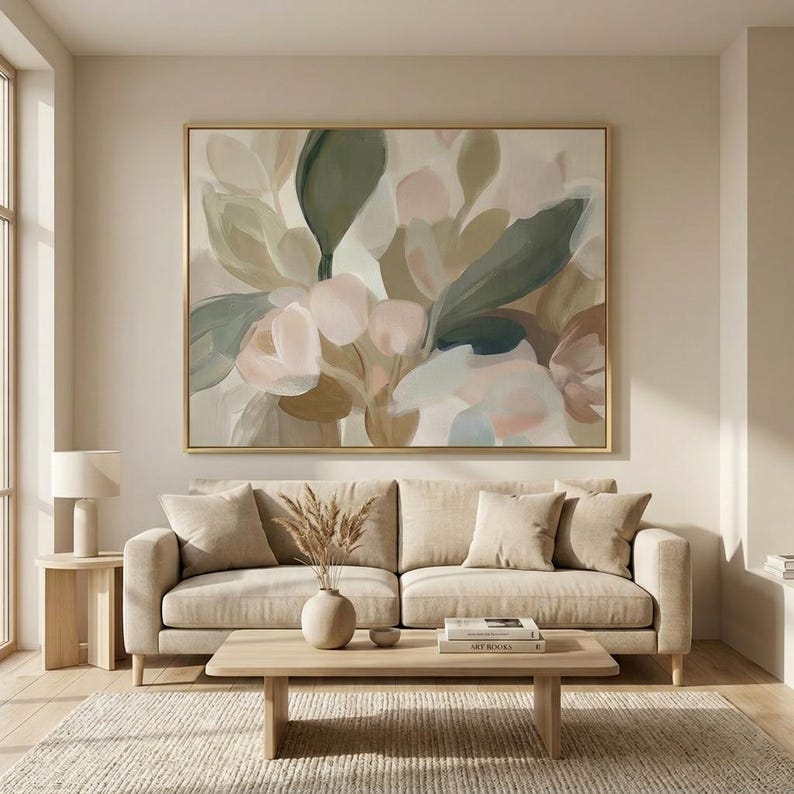 May include: Large abstract floral painting in a gold frame. The artwork features soft colors of green, pink, and beige, creating a calming aesthetic. The painting hangs above a beige sofa with matching pillows.