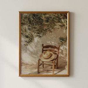 May include: A framed print depicting a wooden chair with a straw hat, positioned against a white wall with olive tree branches. The image has a warm, neutral colour palette, evoking a sense of calm.