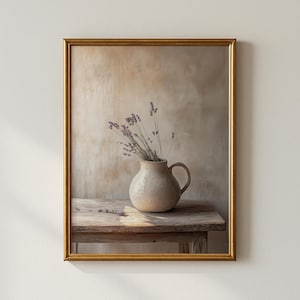 Puede incluir: Impresión enmarcada que presenta una naturaleza muerta. Una jarra de cerámica beige con asa contiene tallos de lavanda seca. La jarra está sobre una mesa de madera rústica, con un fondo neutro.