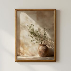 May include: A framed art print featuring a terracotta vase holding olive branches. The artwork has a warm, neutral color palette with a gold frame. The background has a textured, beige wall with shadows.