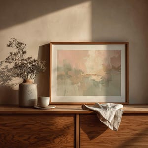 May include: A framed abstract painting with a wooden frame, featuring soft pink, beige, and green hues. A ceramic vase with dried flowers, a small cup and saucer, and a folded linen cloth rest on a wooden cabinet.
