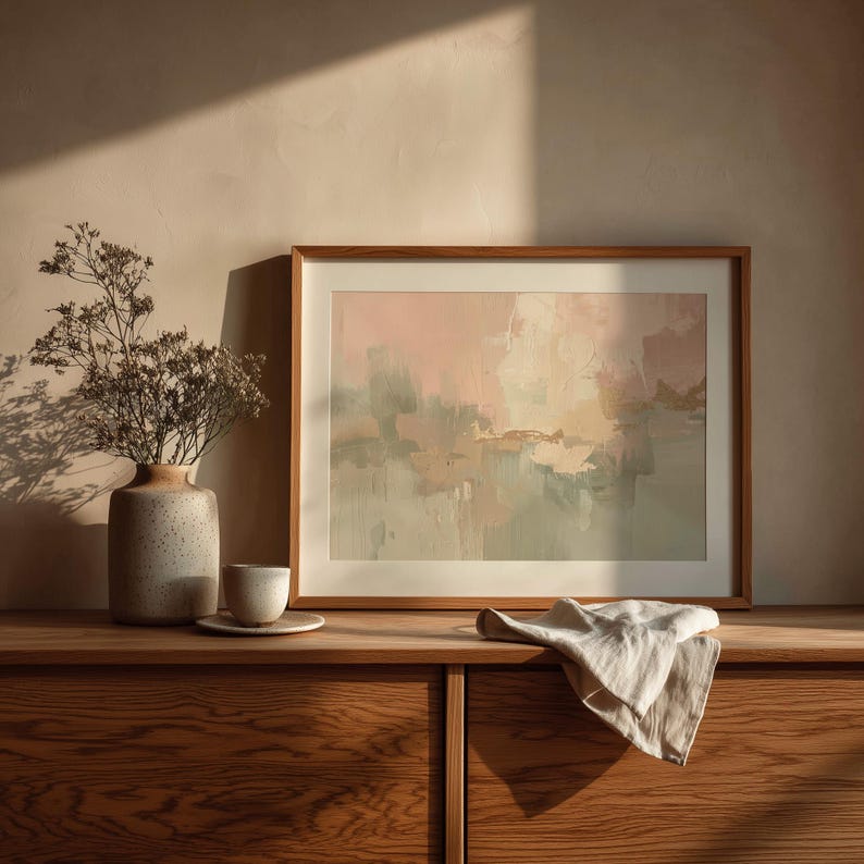 May include: A framed abstract painting with a wooden frame, featuring soft pink, beige, and green hues. A ceramic vase with dried flowers, a small cup and saucer, and a folded linen cloth rest on a wooden cabinet.