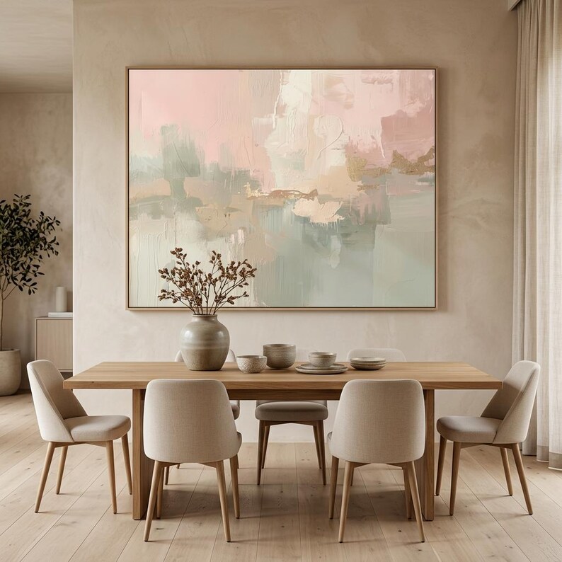 May include: A dining room scene featuring a large abstract painting in soft pinks, greens, and gold tones. A wooden dining table is surrounded by beige upholstered chairs. A vase with dried branches sits on the table.