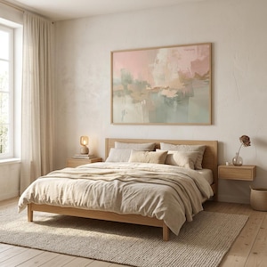 May include: A bedroom scene featuring a wooden bed frame with beige bedding, pillows, and a blanket. A large abstract painting in shades of pink, green, and beige hangs above the bed. The room includes a bedside table, a lamp, and a woven rug.
